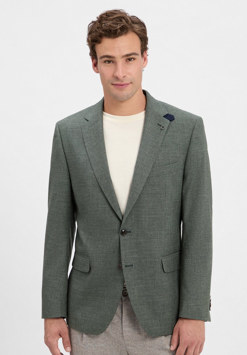 Junger Mann in einem hellgrünen Blazer über einem elfenbeinfarbenen Hemd, mit lockigem Haar und einem leichten Lächeln, posiert vor einem schlichten weißen Hintergrund.