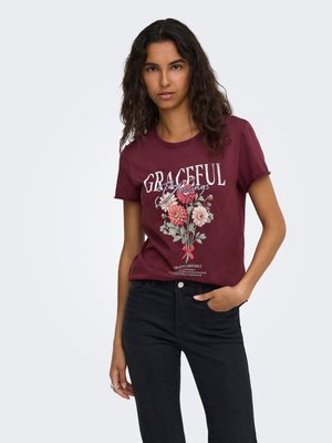 Joven con cabello oscuro y ondulado lleva una camiseta granate con diseño floral y texto "Graceful Blessings" combinada con vaqueros negros.