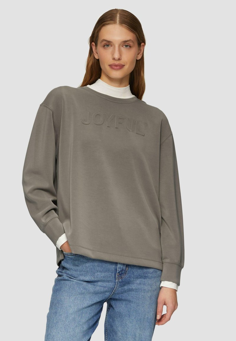 Grijze oversized sweatshirt gemaakt van zachte stof, met een geribde halslijn en reliëf tekst "JOYFUL" aan de voorkant. Gecombineerd met blauwe denim jeans.