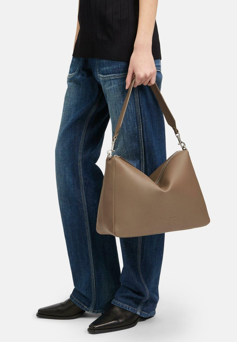 Brązowa skórzana torba na ramię w kształcie trójkąta, z zamkiem błyskawicznym i odpinanym paskiem. Model nosi luźne niebieskie dżinsy.