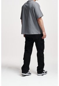 Szara, oversize'owa koszulka, wykonana z bawełny, z krótkimi rękawami. Czarne spodnie cargo z bocznymi kieszeniami. Czarno-białe sneakersy. Gładkie tło.