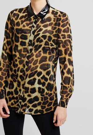 Transparante blouse met lange mouwen en luipaardprint, met zwarte glanzende puntkraag en zwarte knopen, gedragen met zwarte broek.
