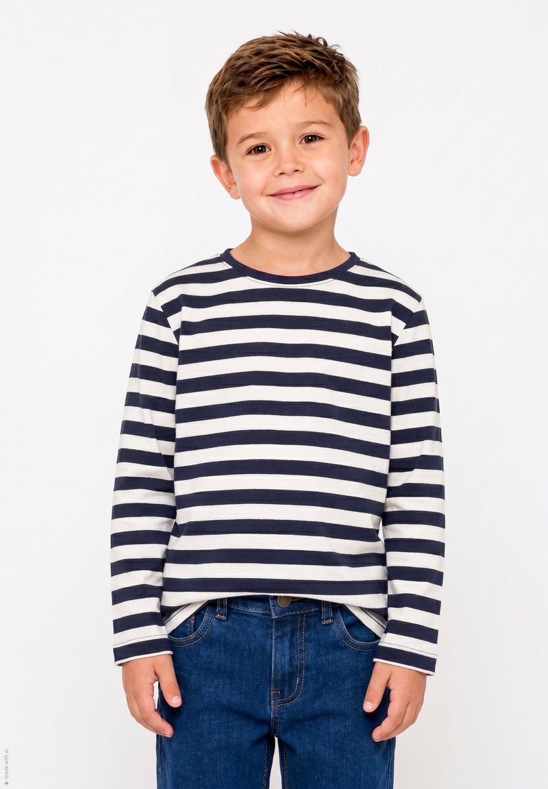 Jeune garçon souriant portant un t-shirt à manches longues rayé bleu marine et blanc avec un jean bleu, debout devant un fond blanc uni.