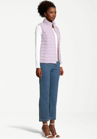 Chaleco acolchado morado claro con cuello alto, combinado con una camisa blanca de manga larga y pantalones de mezclilla. Modelo que lleva sandalias de tacón blancas.