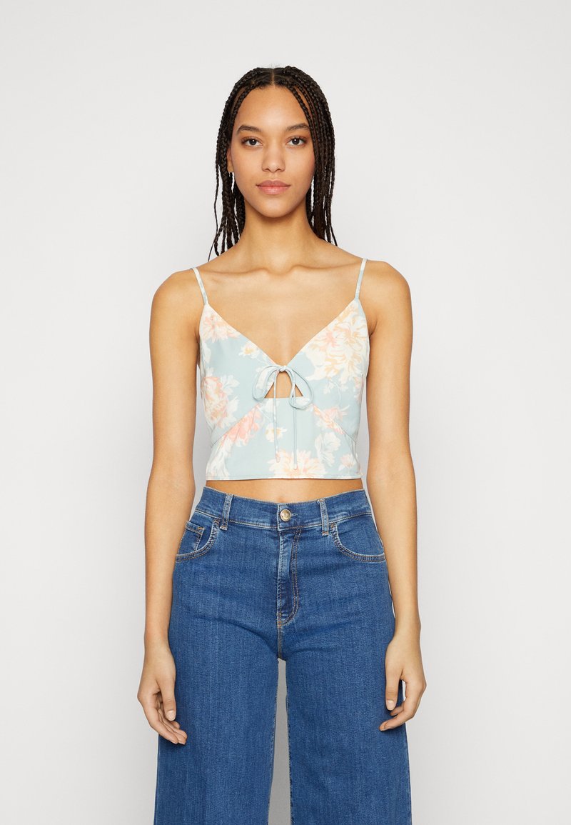 Top cropped floral em azul claro com flores em rosa-laranja, alças spaghetti e um laço na parte da frente. Combinado com jeans azuis de cintura alta.