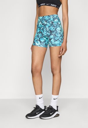 Vrouw draagt een zwarte Nike sportbeha, blauwe patroon korte broek met hoge taille, witte Nike sokken en zwarte Nike sneakers, staand tegen een effen achtergrond.
