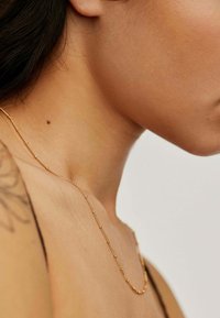 Collar de cadena de oro delgada con pequeñas cuentas redondas y ovaladas, cayendo elegantemente a lo largo del cuello. El fondo es simple y discreto.