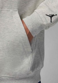 Hand in pocket of light gray sweatshirt sleeve with black Jumpman logo near wrist, showing fabric texture and seam details.