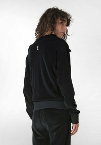 Giacca di velluto nero con colletto alto, polsini a coste e dettaglio del logo sulla schiena. Tessuto morbido e vestibilità rilassata. Abbinata a pantaloni coordinati.