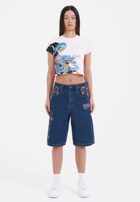 Camiseta gráfica rosa recortada con diseño de dragón azul, combinada con pantalones cortos de mezclilla azul con bordado floral y bolsillos delanteros. Zapatillas blancas.