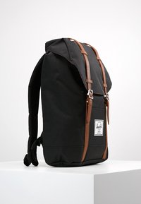 Sac à dos en toile noire avec des sangles et des accents en cuir marron, doté d'un rabat supérieur et d'un patch de marque à l'avant. Panneau arrière texturé.