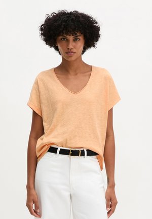 Vrouw met krullend haar die een perzikkleurig T-shirt met V-hals en korte mouwen draagt, ingestopt in een witte broek met een zwarte riem, staand tegen een effen achtergrond.