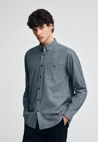 Skjorta med knäppning framtill i mörkgrått tyg, med långa ärmar, en bröstficka och svarta knappar. Diskret struktur och logotyp på fickan.