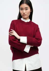 Burgundské svetr s vroubkovanými manžetami a lemem, přesažený přes bílou košilovou košili. Měkká textura a pohodlný střih.