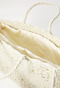 Austinas tamborā maku, kas izgatavots smalkā baltā krāsā ar teksturētu, sīki izstrādātu rakstu. Apļveida malas, divas pītās rokturi un smilšu krāsas auduma odere iekšpusē.
