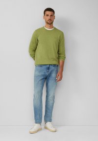 Grünem leichten Rundhals-Pullover, blauen Jeans und weißen Sneakern, vor einem einfachen grauen Hintergrund stehend. Minimalistisches Design, lässige Passform.