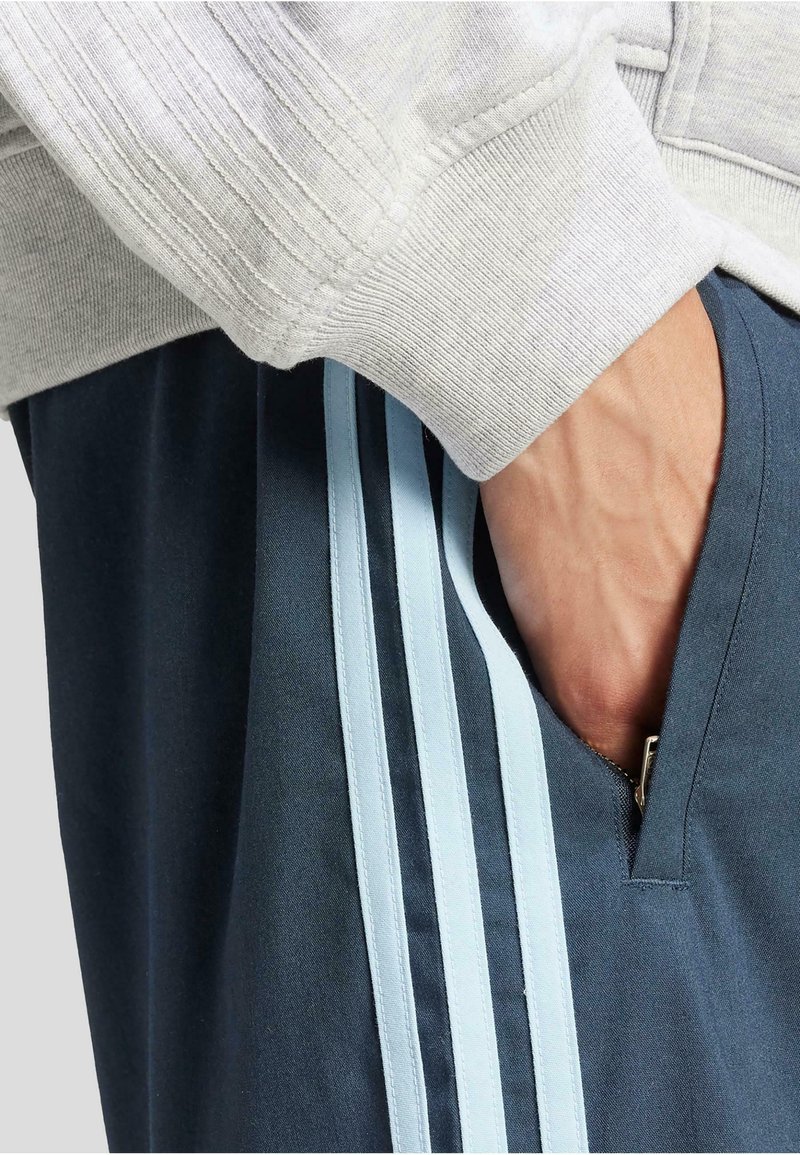 Manche grise côtelée avec un pantalon de jogging bleu marine présentant des rayures latérales bleu pâle et une poche zippée. Main insérée dans la poche, révélant un motif texturé.