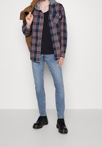 Camisa de franela a cuadros en azul marino, rojo y amarillo con forro de sudadera gris; camiseta negra, jeans azul claro y zapatos negros con costuras amarillas.