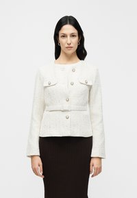 Veste en tweed blanc à la coupe structurée, dotée de deux poches avant, de boutons dorés et d'une ceinture à la taille. Manches longues légèrement évasées.