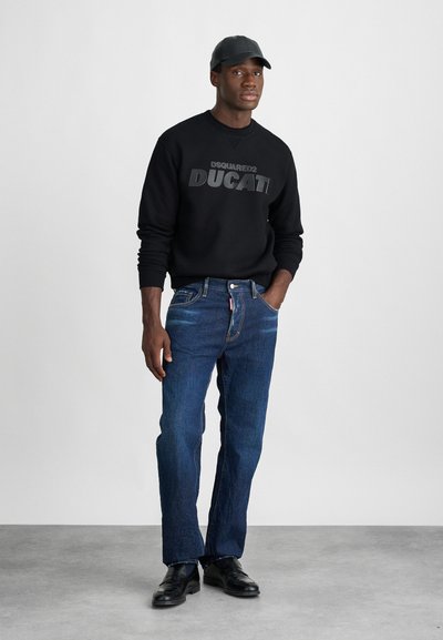 Zwarte sweatshirt met het "DSQUARED2 DUCATI" logo, gecombineerd met blauwe jeans en zwarte schoenen. Model draagt een zwarte pet. Eenvoudige achtergrond.