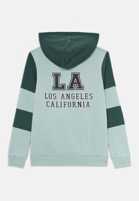 Cappucino felső világos türkiz színben, sötétzöld részletekkel. A hátsó részén "LA" és "LOS ANGELES CALIFORNIA" felirat található. Pamut keverék anyag.