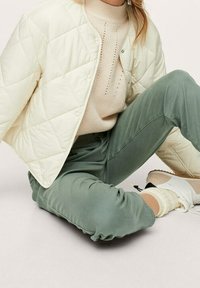 Giacca trapuntata color crema, maglione beige a coste e pantaloni verdi a sigaretta. Sneakers bianche e calzini crema visibili. Texture morbide, vestibilità casual.
