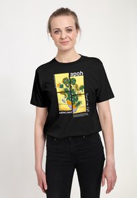 Czarny t-shirt z nadrukiem przedstawiającym słoneczniki w wazonie na żółtym tle, z elementami tekstowymi i kwadratową ramką.