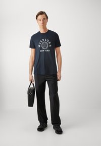 Tricou din bumbac de culoare albastru marin, cu un logo grafic alb, asortat cu pantaloni negri și adidași negri. Utilizatorul ține o geantă de mână neagră.