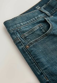 Jean en denim bleu plié avec poche avant visible, petite poche à monnaie, rivet en cuivre et étiquette de taille sur la ceinture.