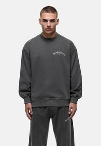Grijze sweatshirt met ronde hals en verlaagde schouders, ribgebreide manchetten en onderkant, voorzien van witte cursieve tekst op de borst. Losse pasvorm, zachte textuur.