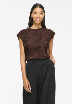 Femme souriante aux longs cheveux foncés portant un haut marron sans manches à motif floral et un pantalon noir taille haute, debout avec une main dans la poche.