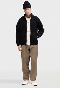 Schwarze Fleecejacke mit Reißverschluss und zwei Vordertaschen, getragen über einem weißen T-Shirt und hellbeigen, strukturierten Hosen; dazu eine schwarze Mütze.