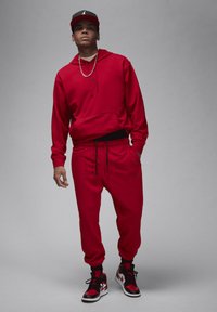 Červená mikina s kapucňou a zodpovedajúce jogger nohavice, s elastickým pásom, šnúrkami a detailom loga. Čierne a červené tenisky dokončujú outfit.