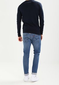 Tmavě modrý pletený svetr a světle modré džínové kalhoty. Džíny mají zadní kapsy s dekorativním prošitím; bílá teniska doplňuje outfit.