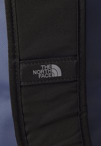 Black fabric strap with a textured surface featuring the logo "THE NORTH FACE" in gray. The background is a smooth blue surface.
