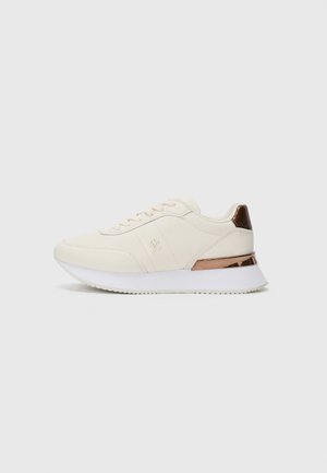 White lace-up sneaker with thick white sole and metallic bronze heel accent, side logo embossed on soft textured upper.