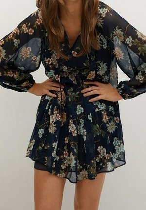 Femme portant une robe fleurie sombre et transparente à manches longues, mains sur les hanches, et une jupe courte à volants.