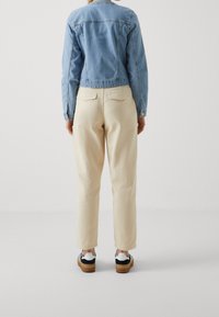 Persoon die met de rug naar ons toe staat, gekleed in een lichtblauwe spijkerjack, crèmekleurige broek met achterzakken, en zwart-witte sneakers.