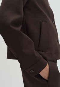 Bruine jas met een gestructureerd ontwerp, voorzien van een zijzak, met knopen gesloten manchetten en een gladde textuur van de stof.