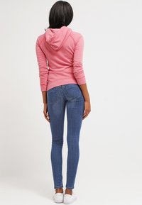 Sudadera rosa con cordón, puños y dobladillo acanalados; combinada con jeans ajustados azules y zapatillas blancas, vista desde atrás.