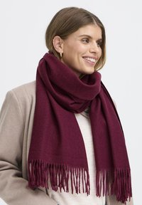 Bordová šatka z mäkkej látky s strapcovými okrajmi. Voľne nosená okolo krku, kombinovaná s svetlo béžovým kardigánom.