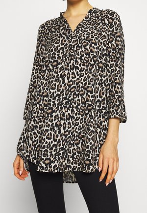 Vrouw draagt een losse, lange mouw luipaardprint tuniek met een V-hals, gecombineerd met een zwarte broek, tegen een effen achtergrond.