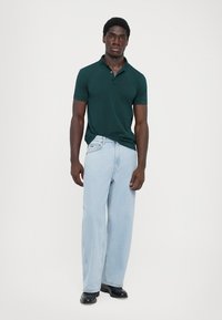 Polo shirt verde închis cu un logo subtil, asortat cu blugi evazați de culoare albastru deschis. Modelul poartă pantofi negri, stând pe un fundal simplu.