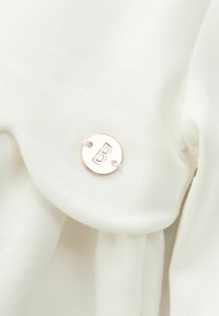 Bílá látka s texturovaným povrchem, na které je připevněna kruhová kovová cedulka s vyrytým písmenem „B“ v moderním písmu a růžově zlaté barvě.