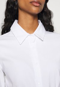 Camicia bianca con colletto a punta, tessuto liscio e cinque bottoni lungo la parte anteriore, con maniche arrotolate e vestibilità rilassata.