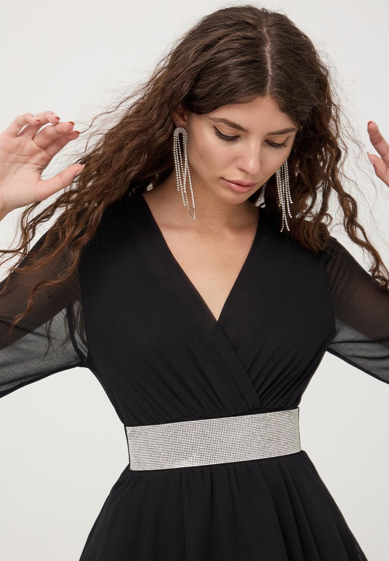 Abito nero con scollatura a V profonda e maniche lunghe trasparenti, dotato di una ampia cintura tempestata di strass argento. I capelli sono acconciati in onde morbide.