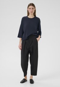 Marineblauer Strickpullover mit weitem Ärmel, kombiniert mit schwarzen, plissierten Hosen und schwarzen Slippern, vor einem schlichten weißen Hintergrund.