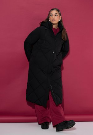 Mujer con un abrigo negro acolchado largo, pantalones rojos de pierna ancha y botas negras, de pie contra un fondo rojo mirando de lado.