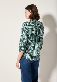 Camicia a fantasia verde menta con disegni geometrici neri, caratterizzata da maniche a tre quarti e una vestibilità rilassata. Dettaglio con piega sul retro visibile.