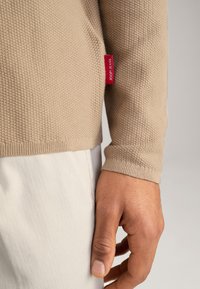 Pull en beige texturé à manches longues, avec une étiquette rouge Joop! Jeans sur le côté. Assorti avec un pantalon de couleur crème.